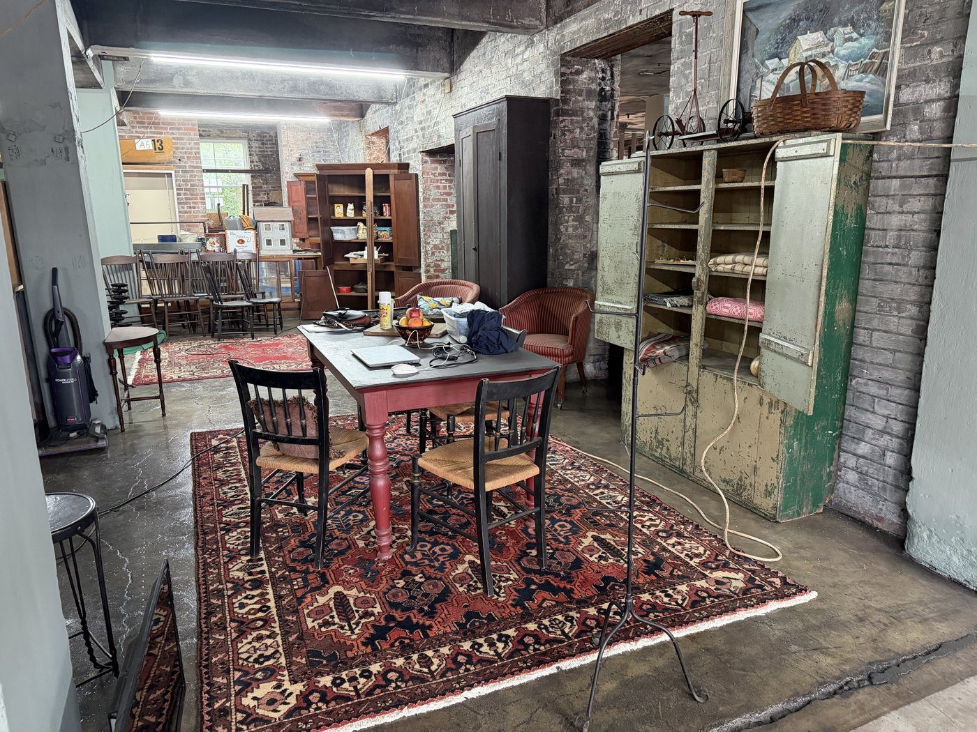 A painted farm table and rush-seat chairs on an oriental rug, with an old green cupboard against whitewashed brick