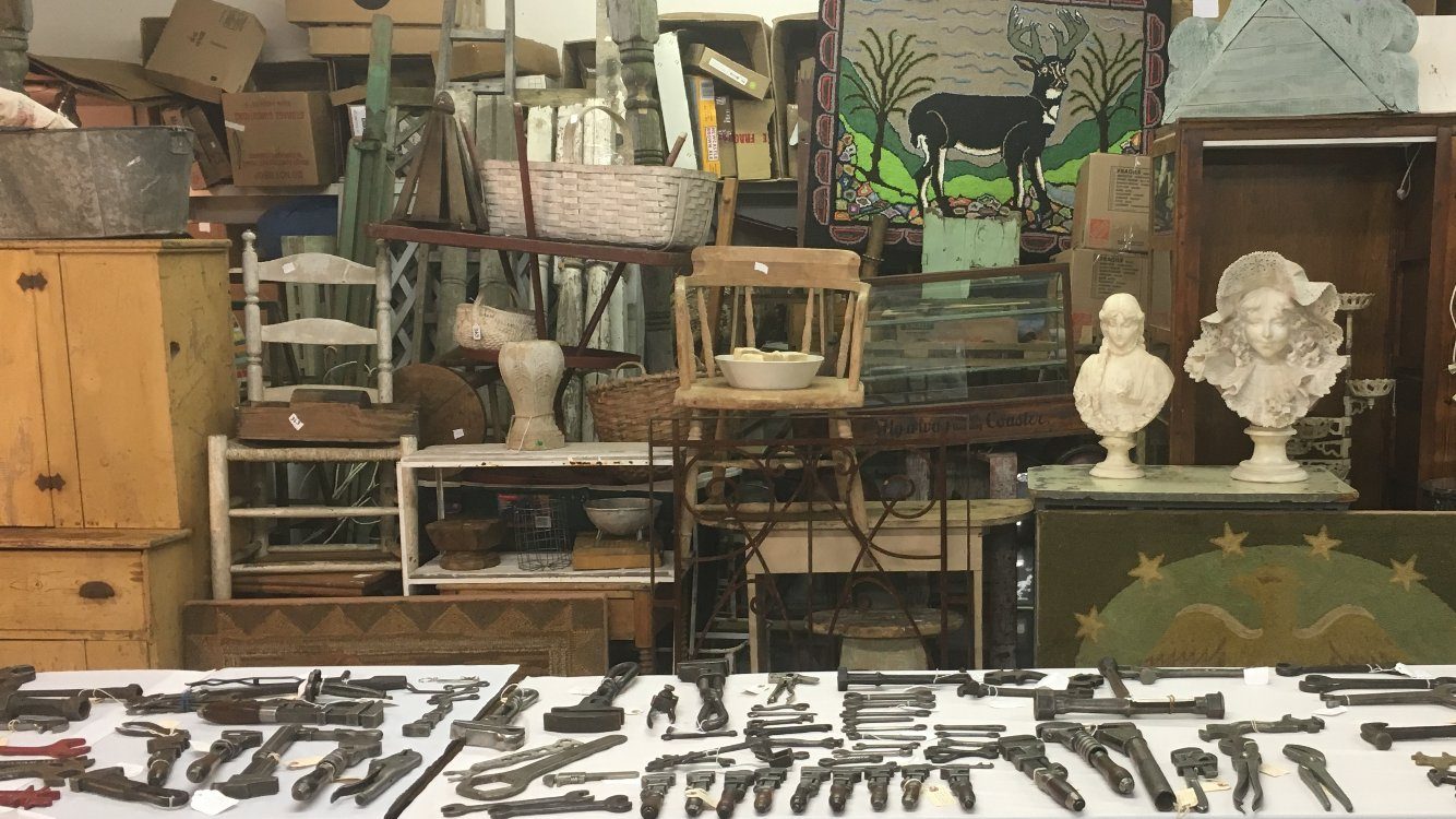 A table of vintage wrenches and tools in front of primitive furniture, a hooked deer rug and marble busts
